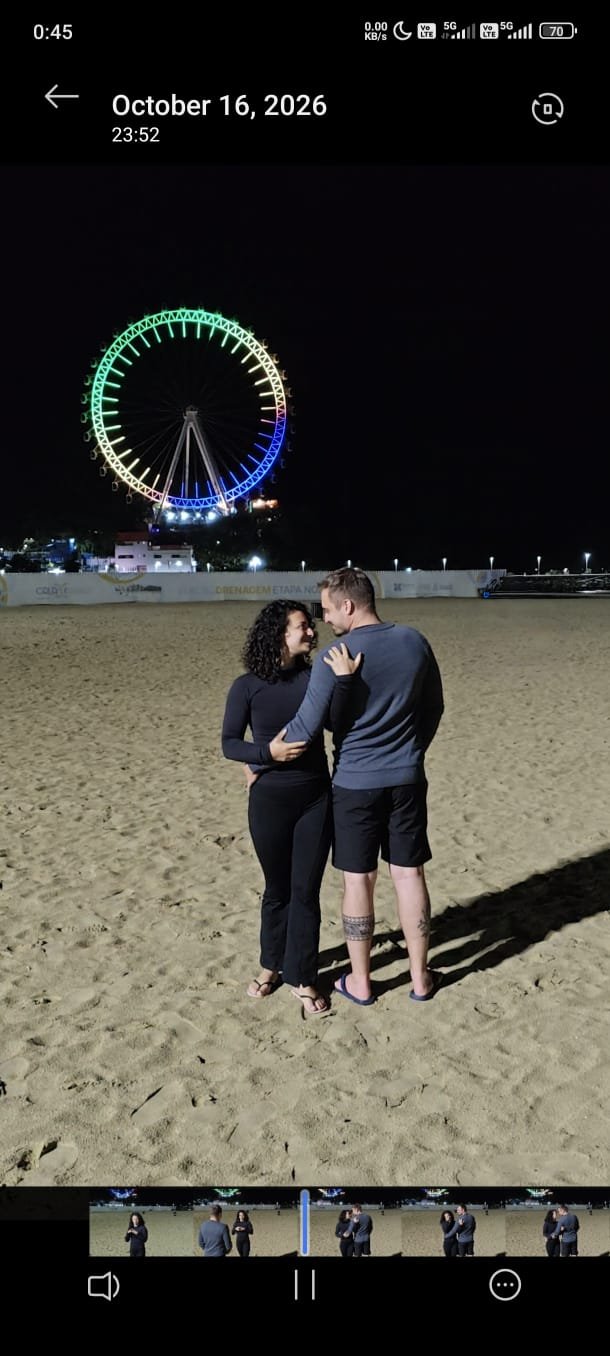 Lara e Rafael abraçados na praia, com a roda gigante iluminada ao fundo